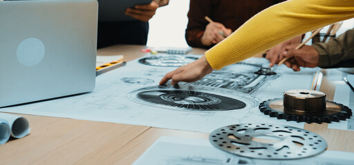 A diverse team collaborates around a table filled with technical drawings and design tools. Focus on hands working on engineering concepts in a modern office setting. SACTR