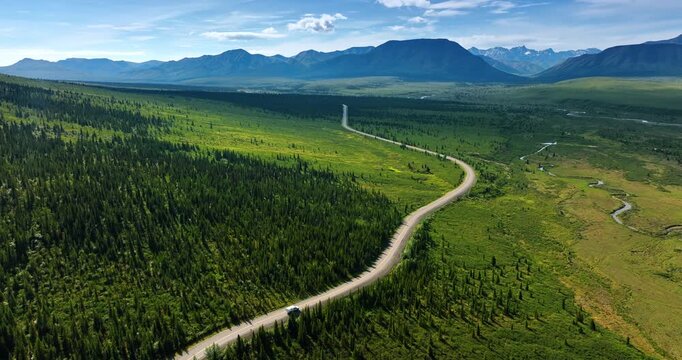 Campervan rides by the wavy road crossing the green valley. Spectacular mountain range at backdrop. Alaska wilderness. Aerial view.