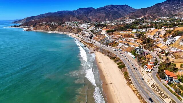 Numerous cars ride by the highway along the sandy beach of the Pacific Ocean. Drone flight along the rocky coast of Malibu, Los Angeles County, California, USA.