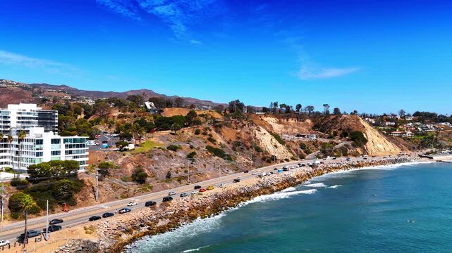 Lively road with many cars parked along the rocky shore of the Pacific Ocean. Aerial perspective on sunny Malibu, Los Angeles County, California, USA.