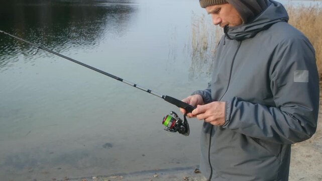 Adult male angler in a warm jacket assembling a professional spinning fishing rod, preparing his tackle and equipment for fishing on the calm lake shore during a cool autumn day