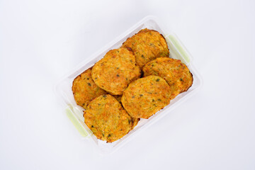 A close-up image of homemade vegetable fritters, neatly arranged in a transparent plastic container. The golden-brown patties appear crispy on the outside with visible bits of herbs and vegetables.
