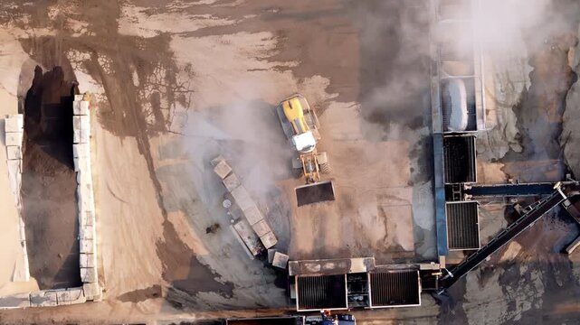 Aerial top-down view showing a front loader transferring sand and gravel into a sorting or crushing machine. Dust rises as the heavy machinery operating