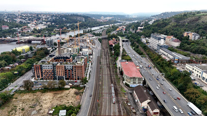 Aerial view of cityscape with parallel railway tracks and busy highway beside river. Urban contrast between movement, architecture, and industrial landscape captured from above.
