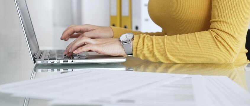 Woman focusing on her work, typing on a laptop keyboard, sitting at a glass desk with paperwork and folders, managing digital and administrative tasks in a modern office environment