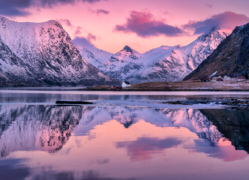 Fototapeta Beautiful pink sunrise illuminating snowy mountains and houses reflecting in the calm water of a fjord in Lofoten islands, Norway. Winter landscape with sea, pink sky with clouds, rorbu, rocks in snow
