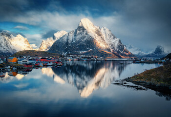 Snowy mountains and colorful houses reflect in a fjord under dramatic clouds at sunset. Winter...