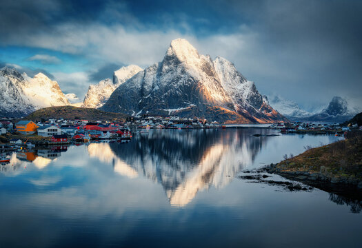 Fototapeta Snowy mountains and colorful houses reflect in a fjord under dramatic clouds at sunset. Winter scenery with sea, rocks in snow, cloudy blue sky, rorbu. Reine village in Lofoten, Norway. Landscape