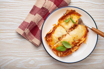 Lasagna with rich bolognese sauce, melted cheese, and fresh basil on a white plate, served on white plate on a light wooden background with a checkered napkin and fork, copy space