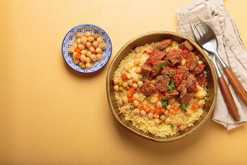 Traditional dish of Maghreb cuisine, a Moroccan couscous with lamb or beef, vegetables, chickpeas, and peas in a rustic bowl with cutlery on yellow background, copy space