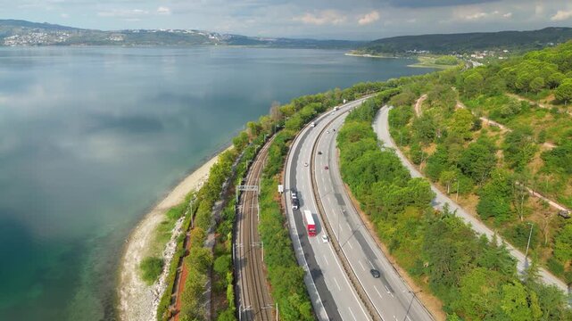 Aerial view of a high-speed train in motion, speeding past a highway and a lakeside promenade at Sapanca Lake. Modern transportation infrastructure and nature coexisting.