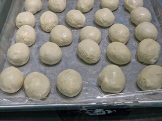 High-angle view of several rows of small, round balls of raw dough arranged closely on a metal baking sheet that is partially covered with a clear plastic sheet.