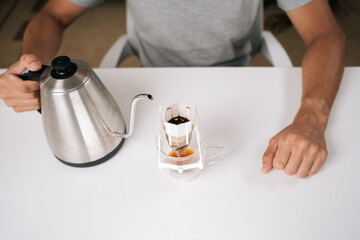 Cropped shot of person brewing drip coffee with kettle, carefully pouring hot water over paper filter resting on transparent glass cup, all set on clean white table in cozy kitchen.