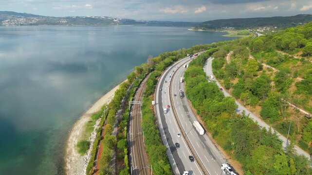 Aerial view of modern infrastructure and nature coexisting at Sapanca Lake: Static drone shot capturing a high-speed train, motorway, and pedestrian promenade side-by-side on the lake shore.