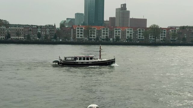 A long cargo container barge travels along the Nieuwe Maas river in Rotterdam, Netherlands, against a cloudy sky. The vessel carries international shipping containers past the impressive city skyline 