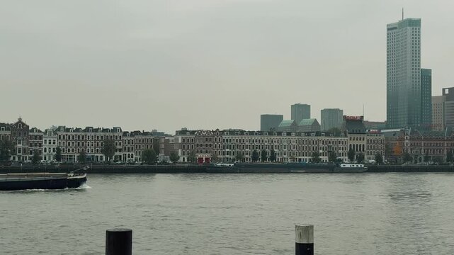 A long cargo container barge travels along the Nieuwe Maas river in Rotterdam, Netherlands, against a cloudy sky. The vessel carries international shipping containers past the impressive city skyline 
