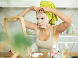 Adult woman in towel turban on head applying hydrating face mask during daily self-care routine...