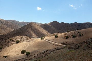 Arid rolling hills of southern Crete under a bright blue sky, with sparse vegetation, dirt road, and warm sunlight highlighting the rugged Mediterranean terrain.