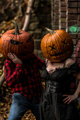 Creative couple dressed for Halloween with carved pumpkin heads poses in a rustic setting surrounded by autumn leaves