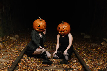 Two friends celebrate autumn with creative pumpkin heads while sitting on a train track covered in fall leaves