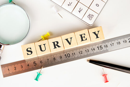 Wooden blocks spelling SURVEY sit next to a ruler. A calculator, magnifying glass, pen, and several push pins are scattered around on a white background