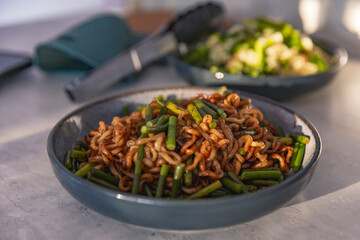 Stir fried noodle dish in ceramic bowl with vibrant scallions, bathed in golden sunlight in a warm kitchen scene with a soft focus background