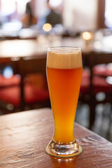Tall glass of wheat beer with thick foamy head standing on wooden bar counter, blurred background of restaurant interior and patrons
