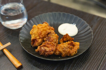 Plate of Japanese karaage fried chicken served with dipping mayonnaise on black plate, chopsticks and water glass on dark wooden table