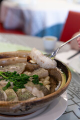 Close up of Chinese fish fillet in clay pot, spoon lifting slice revealing garlic cloves and clear broth with soft texture and herbal garnishing