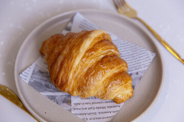 Classic croissant with golden flaky layers and glossy surface, placed on printed paper over ceramic plate, highlighting buttery crust