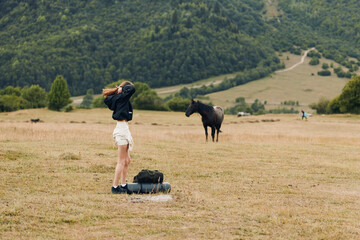 A lone traveler stands in a vast field, backpack down, adjusting her hat as distant horses graze. The expansive landscape conveys adventure, solitude, and outdoor exploration.