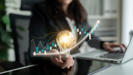 A business professional engages in innovative thinking by holding a glowing light bulb amid financial growth charts, symbolizing creativity and progress. Impute