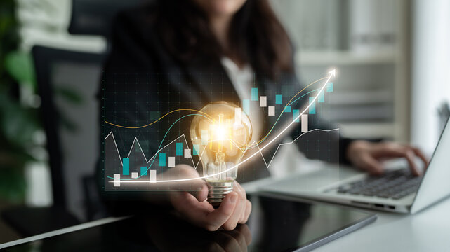 A business professional engages in innovative thinking by holding a glowing light bulb amid financial growth charts, symbolizing creativity and progress. Impute