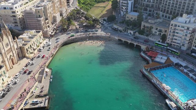 Late-afternoon 4K drone over Balluta Bay, Malta capturing turquoise bay, beach swimmers, rooftop pool, Gothic church, coastal promenade and urban seaside life for travel, real estate, lifestyle use