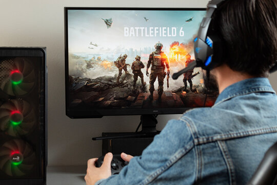 a male gamer at the right side of the picture holding a xbox controller watching a pc monitor with battlefield 6 shooter video game