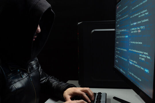 a hooded man writing on a keyboard computer in front of computer monitor with blue html code - Powered by Adobe