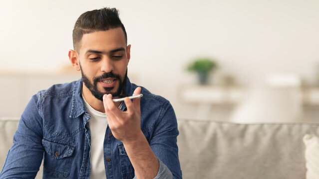 A man with a beard interacts with his smartphone using voice command. He sits on a couch in a stylish living room filled with comfortable decor and plants.