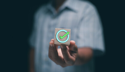 Concept of approval and successful completion. Hand holding wooden cube with glowing green check mark. Symbolizing validation, achievement, confirmation and business success in digital technology. © krungchingpixs