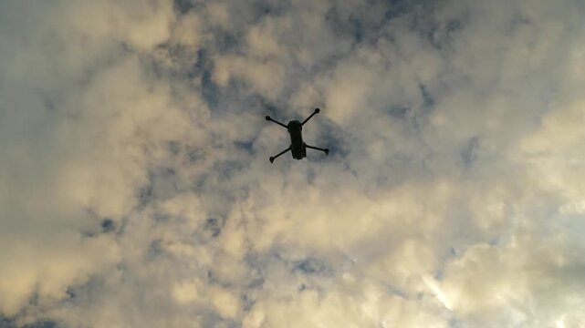 Under-view of hovering quadcopter stationary against clouded sky