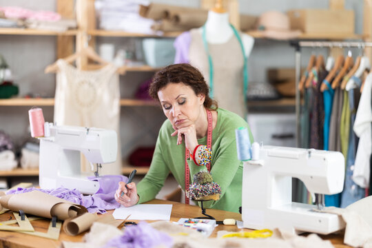 Female clothing designer sitting with pen in hand over blank sheet at cluttered worktable, looking sad and deep in thought, troubled by stalled project, feeling creative block and lack of ideas