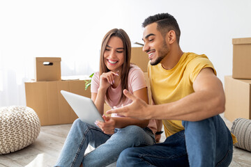 A young couple sits on the floor of their new home, smiling as they look at a tablet together. Cardboard boxes are stacked in the background, suggesting they are relocating.