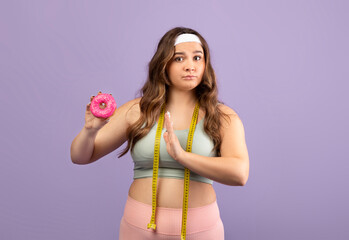 A serious caucasian plus size young woman in sportswear holds a donut and gestures refusal. She...