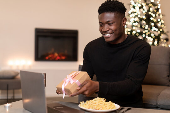 A joyful African American man is video calling while holding a wrapped gift, wishing someone special during a celebration. The warm home setting features a lit fireplace and festive decorations.