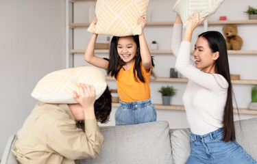 A boy sits on the couch covering his head with a pillow while two girls laugh and swing their own pillows, creating joy in a bright living room setting.