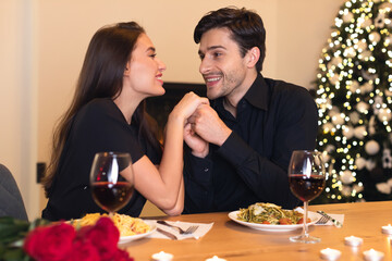 A couple shares a joyful moment during a romantic dinner. They are seated at a table with pasta and wine, surrounded by a festive Christmas tree and candles.