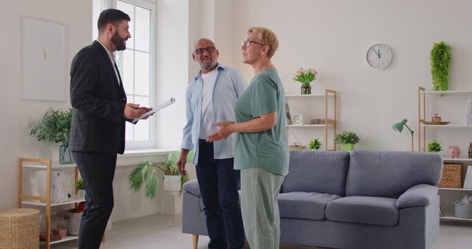 Realtor leads elderly clients on an apartment tour, discussing contract terms, purchase details in a bright living room. Real estate agent guides senior couple through apartment viewing.