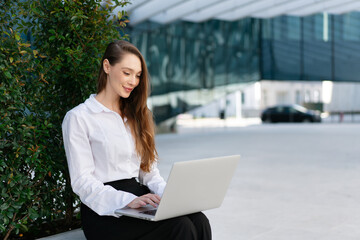Smiling young businesswoman using laptop while working on project. Happy young woman in white shirt sitting near plants with blurred office building in background