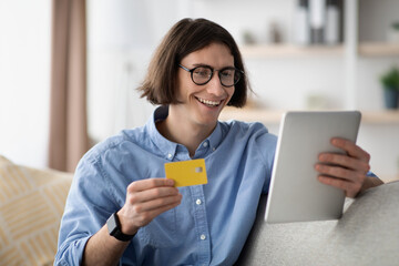A cheerful man sits on a couch in a bright living room. He holds a yellow credit card in one hand while using a tablet with the other, enjoying the online shopping experience.