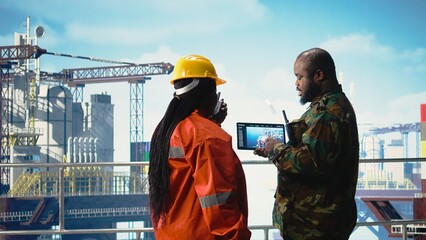 Soldier on offshore platform deck talking with technician, using device visualization software. African american naval unit guards drilling rig against terrorist threats using tablet app, © DC Studio