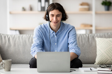 A young man wearing headphones interacts on a video call from his cozy living room. He sits on a sofa with a laptop, enjoying the conversation in a bright setting.